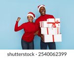 A happy couple dressed in matching red outfits and Santa hats beams with excitement as they hold stacks of present-wrapped boxes decorated with ribbons. The vibrant blue backdrop adds festive cheer.