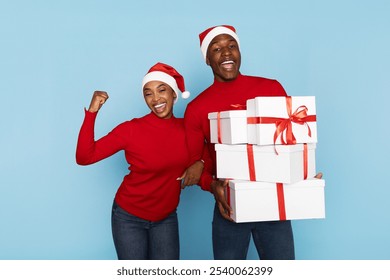 A happy couple dressed in matching red outfits and Santa hats beams with excitement as they hold stacks of present-wrapped boxes decorated with ribbons. The vibrant blue backdrop adds festive cheer. - Powered by Shutterstock