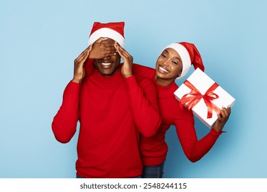 A happy couple dressed in red sweaters and Santa hats enjoys a festive moment. One partner playfully covers the other eyes while holding a wrapped gift, showcasing holiday cheer. - Powered by Shutterstock