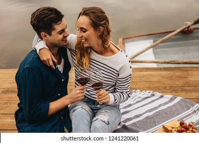 Happy Couple Drinking Wine Sitting Near A Lake And Laughing Holding Each Other. Couple In Love Sitting On A Wooden Dock Beside A Lake With Wine Glasses In Hand And Snacks By Their Side.