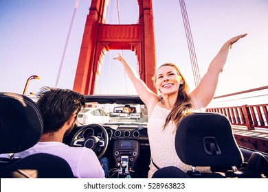 Happy Couple Driving On A Convertible Car - Road Trip To San Francisco, Woman With Raised Arms Greeting Golden Gate Bridge