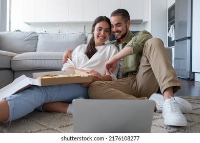 Happy Couple Eating Pizza And Watching Movie. Woman And Man Sitting On Carpet, Holding Pizza, Watching Film From Laptop. Food, Leisure Concept