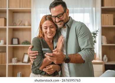 Happy couple embracing while using a smart phone at home, sharing smiles and enjoying a moment of connection through their online experience and digital communication