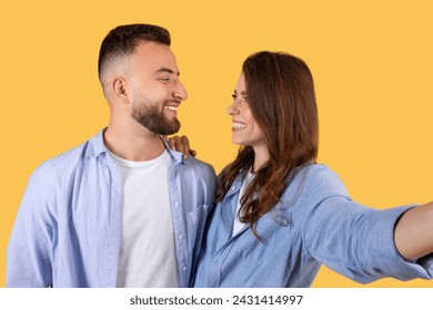 Happy couple engaged in playful moment, taking selfie with genuine smiles as they gaze into each other's eyes, against warm yellow backdrop - Powered by Shutterstock