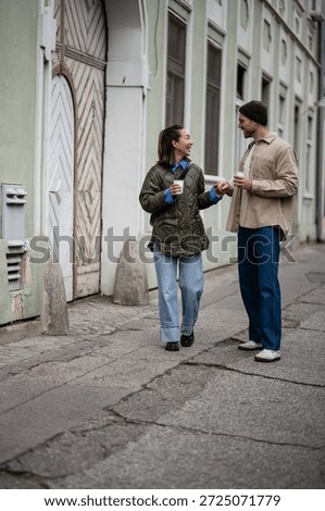Happy couple engaging in conversation while walking city street, holding coffee cups and smiling, enjoying casual lifestyle moment