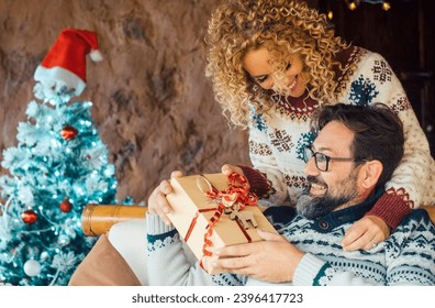 Happy couple enjoy christmas sharing gifts leisure activity at home. Xmas decorations and tree in background. Man receiving a surprise box present from his wife woman behind. People and gift xmas - Shutterstock ID 2396417723