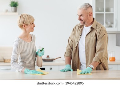 Happy couple enjoying cleaning house together, home hygiene, relationships and domestic life. Happy middle aged husband and wife in protective gloves disinfect table in kitchen interior, empty space - Powered by Shutterstock