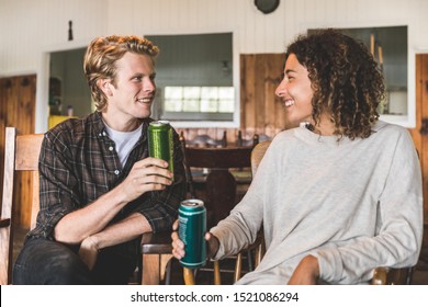 Happy Couple Enjoying A Drink In A Wood Cabin In Canada - Blond Man And Curly Woman Looking Each Other And Smiling, Both Holding A Can Of Beer - Love And Relax Concepts.