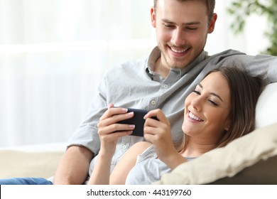 Happy couple enjoying media content in a smart phone sitting on a couch at home - Powered by Shutterstock