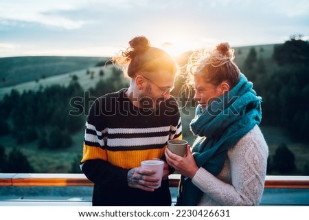 Happy couple enjoying morning coffee and looking at each other while standing on a balcony with an beautiful nature in the background and with a warm light at sunrise. Cold weather.