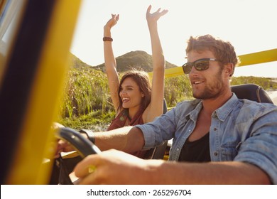 Happy Couple Enjoying On A Long Drive In A Car. Friends Going On Road Trip On Summer Day. Caucasian Young Man Driving A Car And Joyful Woman With Her Arms Raised.