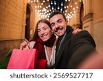 Happy couple enjoying their buying experience with shopping bags while taking a selfie together, capturing a joyful moment filled with excitement and love during a festive evening outing in the city