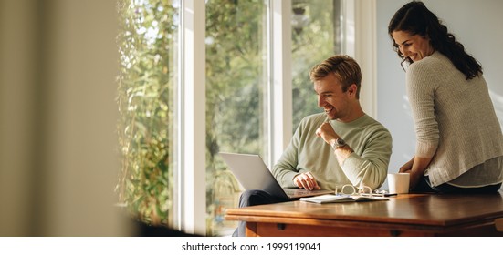 Happy couple enjoying their time at home. Man and woman sitting at tablet looking at laptop. - Shutterstock ID 1999119041