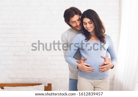 Happy couple expecting baby and hugging near window at home, empty space
