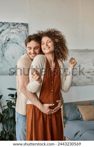 Happy couple expecting baby, man hugging his pregnant wife from behind with cheerful smile