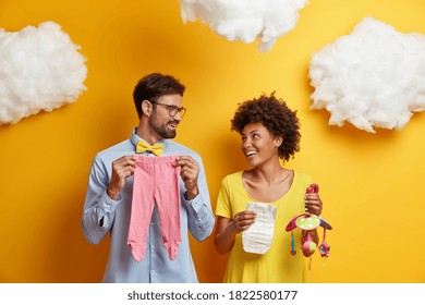 Happy couple future parents prepare for parenthood and giving birth to baby pose with infant items looks cheerfully at each other pose together against yellow wall. Healthy pregnancy family life - Powered by Shutterstock