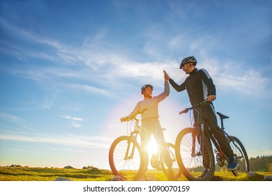 Happy Couple Goes On A Mountain Road In The Woods On Bikes With Helmets Giving Each Other A High Five