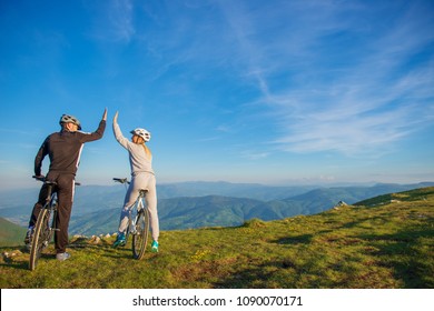 Happy Couple Goes On A Mountain Road In The Woods On Bikes With Helmets Giving Each Other A High Five