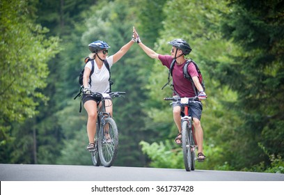 Happy Couple Goes On A Mountain Asphalt Road In The Woods On Bikes With Helmets Giving Each Other A High Five