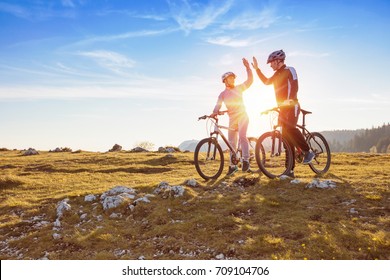 Happy Couple Goes On A Mountain Asphalt Road In The Woods On Bikes With Helmets Giving Each Other A High Five