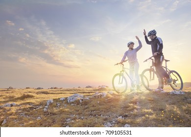 Happy Couple Goes On A Mountain Asphalt Road In The Woods On Bikes With Helmets Giving Each Other A High Five