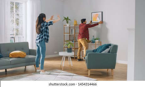 Happy Couple Hanging Picture on the Wall, Boyfriend Moves It, Girlfriend Tells Him when the Frame is Hanging Straight. Funny Moment in Young Couple's Life. Modern Stylish Apartment.