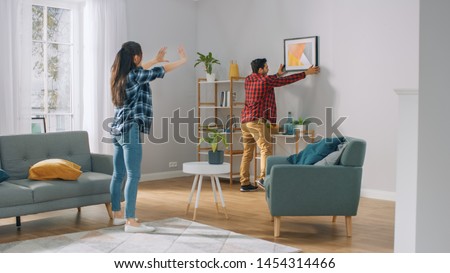 Happy Couple Hanging Picture on the Wall, Boyfriend Moves It, Girlfriend Tells Him when the Frame is Hanging Straight. Funny Moment in Young Couple's Life. Modern Stylish Apartment. 商業照片 © 