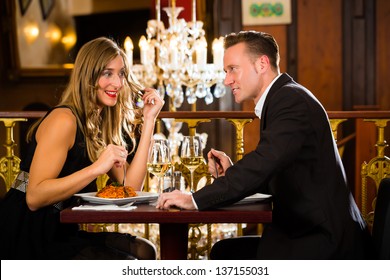 Happy Couple Have A Romantic Date In A Fine Dining Restaurant They Drink Wine And Clinking Glasses, Cheers - A Large Chandelier Is In Background