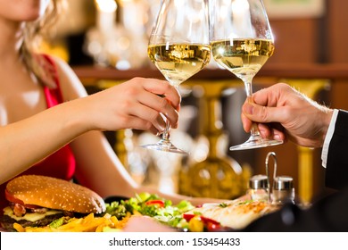 Happy Couple Have A Romantic Date In A Fine Dining Restaurant They Drink Wine And Clinking Glasses, Cheers - A Large Chandelier Is In Background