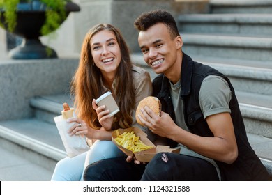 Happy Couple Having A Date In The City. College Students Eating Burger, Potato Frites And Hotdog. Tender Moments In Life.