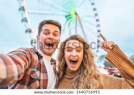 Happy couple having fun at amusement park in London - young couple in love taking a selfie and enjoying time at funfair with rollercoaster on background - Happy lifestyle and love concepts