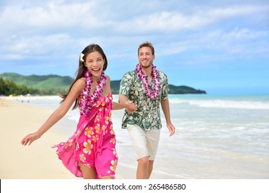 Happy Couple Having Fun Running On Hawaii Beach Vacations In Hawaiian Clothing Wearing Aloha Shirt And Pink Sarong Sun Dress And Flower Leis For Traditional Wedding Or Honeymoon Concept.