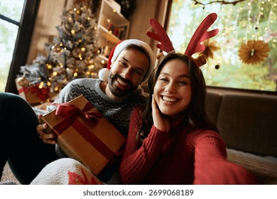 Happy couple having fun while taking selfie on Christmas day at home and looking at camera.