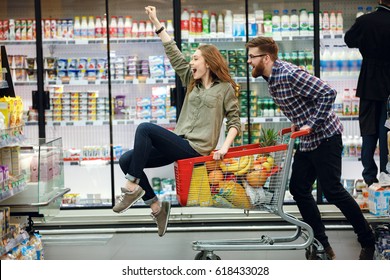Happy couple having fun while choosing food in the supermarket. Young happy man pushing shopping cart with his girfriend inside - Powered by Shutterstock