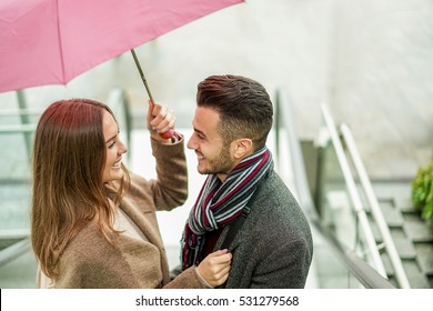 Happy couple having tender moments under a umbrella in rainy day outdoor - Young people smiling going outside of mall shopping center - Love and relationship concept - Focus on woman eye - Warm filter