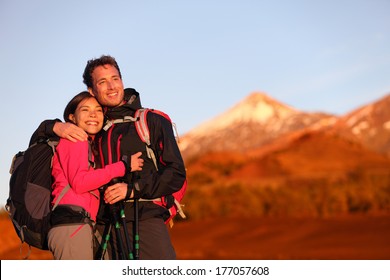 Happy Couple Hiking Enjoying Looking At View Embracing In Love. Hiker Man And Woman Wearing Backpacks Enjoying Sunset During Hike On Mountain Volcano Teide, Tenerife, Canary Islands, Spain.