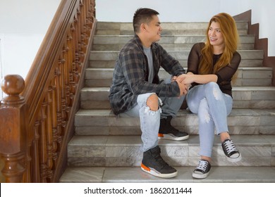 A Happy Couple Hold Hands And Look At Each Other Endearingly While Sitting Down On The Stairs. Concept Of A Strong Relationship And Bond.