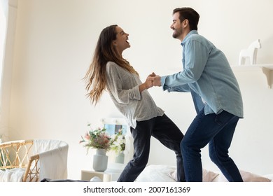Happy couple holding hands laughing having fun fooling together while standing on bed in modern light cozy bedroom. Bank loan, young renters family, romantic relationships and cohabitation concept