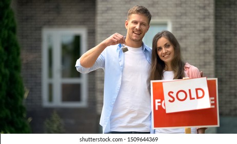 Happy Couple Holding Keys From House Near Sold Sign, Mortgage For Young Families