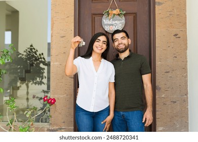 Happy couple holding keys is smiling while standing in front of their new home with a welcome sign - Powered by Shutterstock