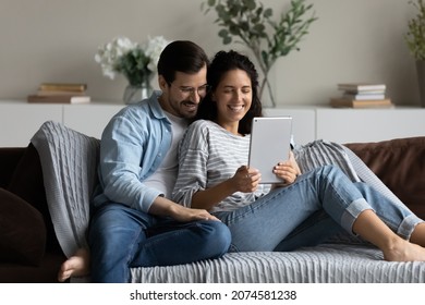 Happy couple hugging, using tablet together, relaxing on couch, smiling beautiful woman and man in glasses looking at screen, chatting online by video call, shopping or watching movie at home - Powered by Shutterstock