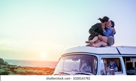 Happy couple kissing on top of minivan roof at sunset - Young people having fun on summer beach vacation - Travel and love concept - Focus on faces - Radial purple and green filters editing - Powered by Shutterstock