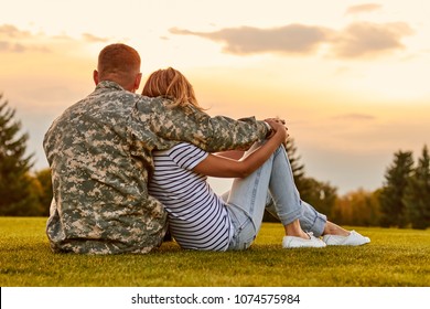 Happy couple looking at sunset. Back view, soldier and his wife sitting on the grass.