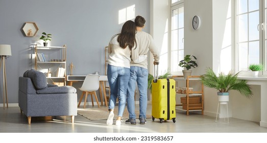 Happy couple in love back home from summer holiday trip. View from behind of young man and woman who have just returned from vacation travel with yellow suitcase standing in living room and hugging - Powered by Shutterstock