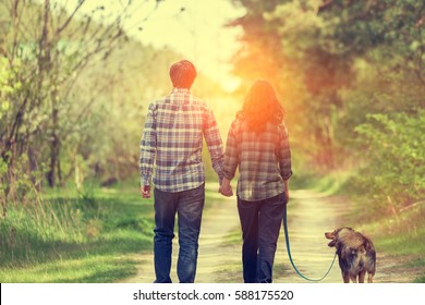 Happy couple in love with dog walking on rural dirt road in springtime at sunset. Woman and man holding hands. Woman keeps her dog on a leash. Couple and dog back to camera. Casual stile. - Powered by Shutterstock