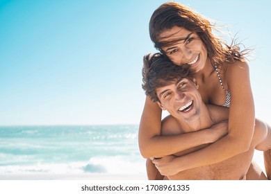 Happy couple in love enjoy summer vacation at beach with copy space. Man giving piggyback ride to his beautiful girlfriend while looking at camera with sea in background. Couple having fun at beach.