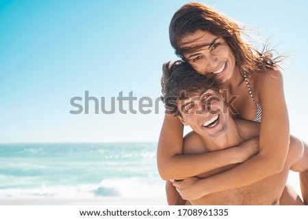Happy couple in love enjoy summer vacation at beach with copy space. Man giving piggyback ride to his beautiful girlfriend while looking at camera with sea in background. Couple having fun at beach.