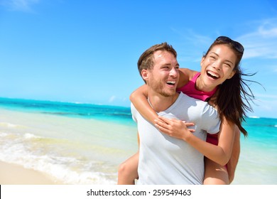 Happy couple in love on beach summer vacations. Joyful Asian girl piggybacking on young Caucasian boyfriend playing and having fun in sunny tropical destination for travel holiday.