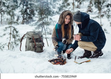 Happy couple in love relaxing in the snow-covered forest roasting marshmallows over bonfire and drinking hot tea - Powered by Shutterstock