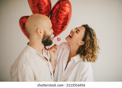 Happy couple in love standing with red heart balloons on white background, celebrating Saint Valentine's Day, laughing and having fun.